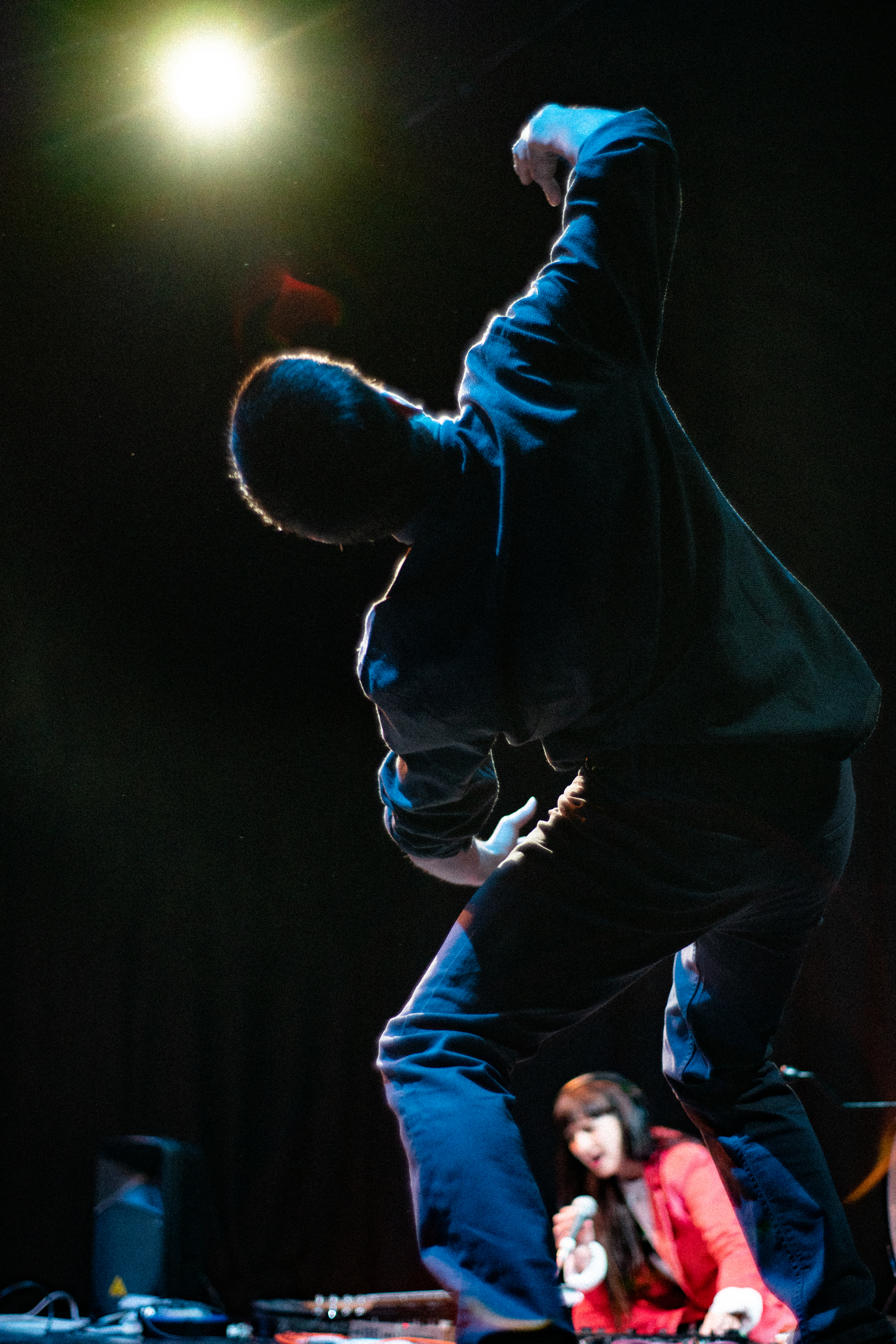 Adam standing on a stage under a spotlight, someone lies still on the floor behind him in the shadow